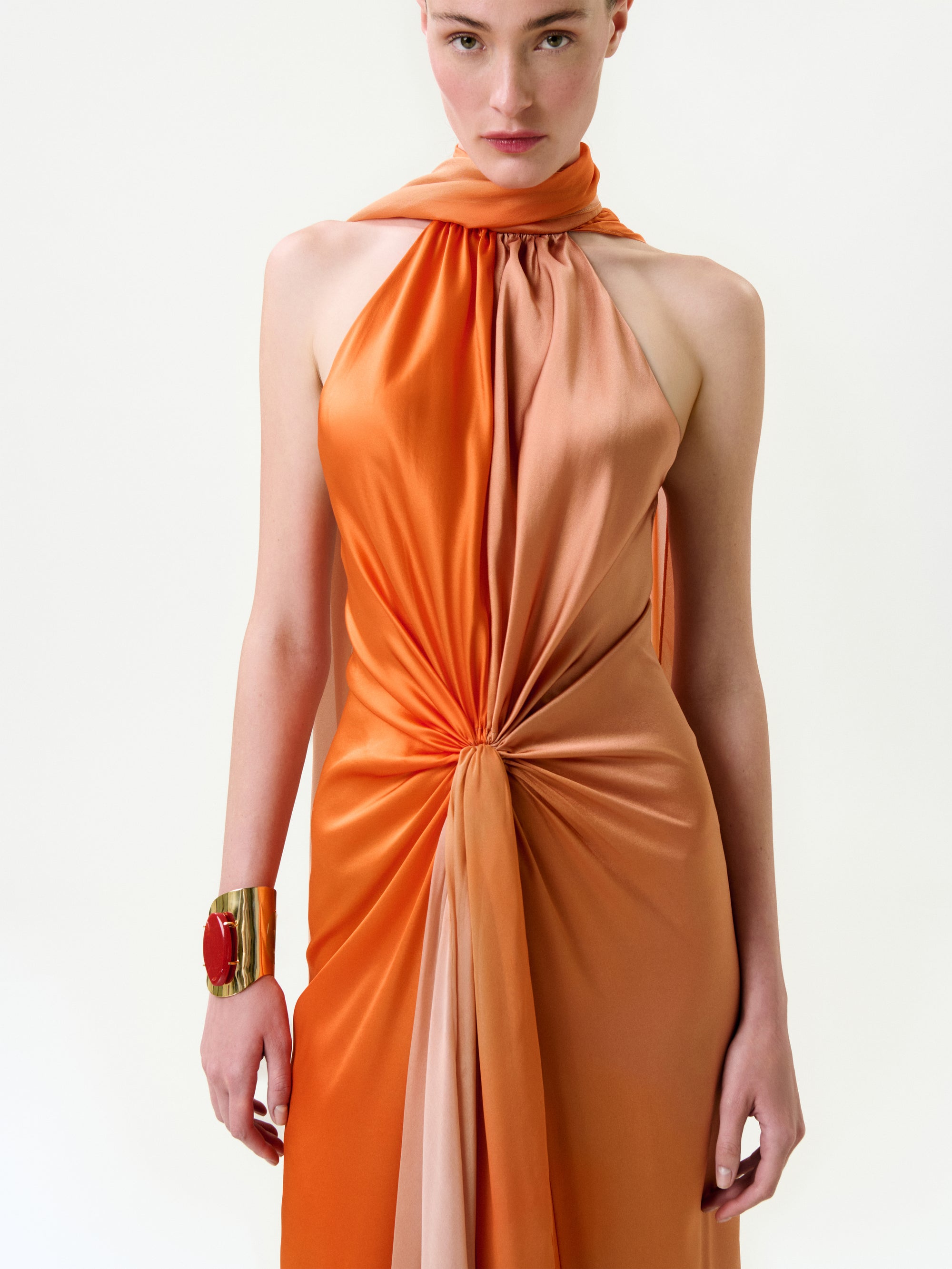 A person wearing the Margalit Dress—a sleeveless, halter-neck silk maxi in orange tones, gathered and knotted at the waist—paired with a large gold cuff bracelet with a red stone, stands against a plain background.