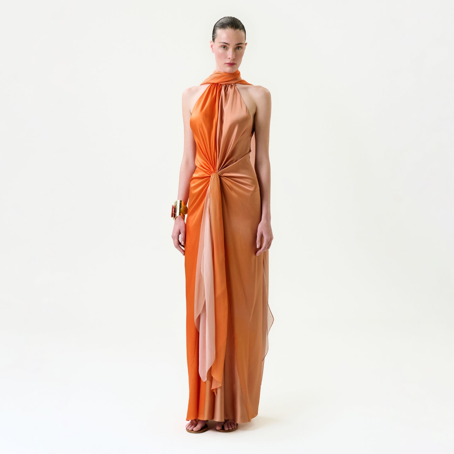 A woman with slicked-back hair and a gold bracelet poses against a white background, wearing the Margalit Dress—a sleeveless, floor-length silk maxi dress in orange and peach with a draped, gathered front.