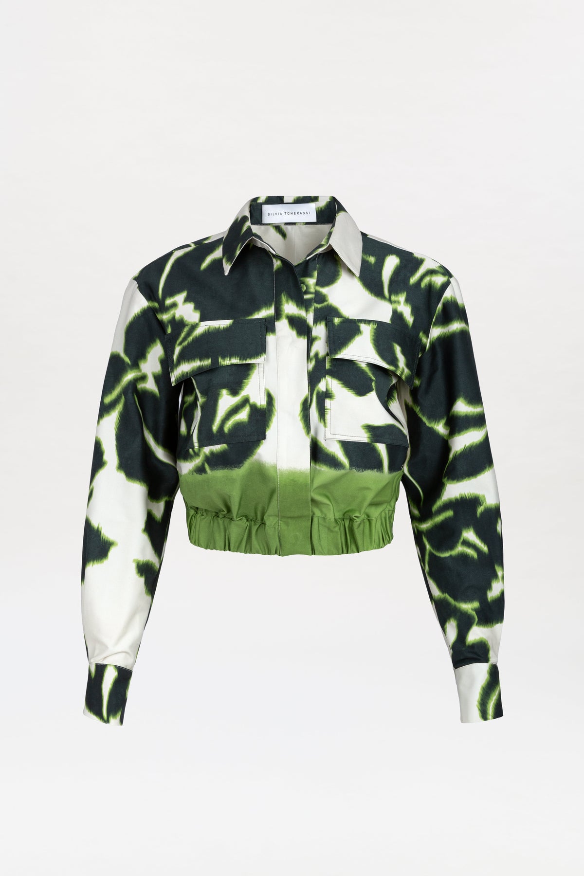 The Soleil Jacket is a cropped jacket featuring a green and black tie-dye pattern, long sleeves, a collar, a front pocket, and a green elastic waistband, displayed on a plain white background.