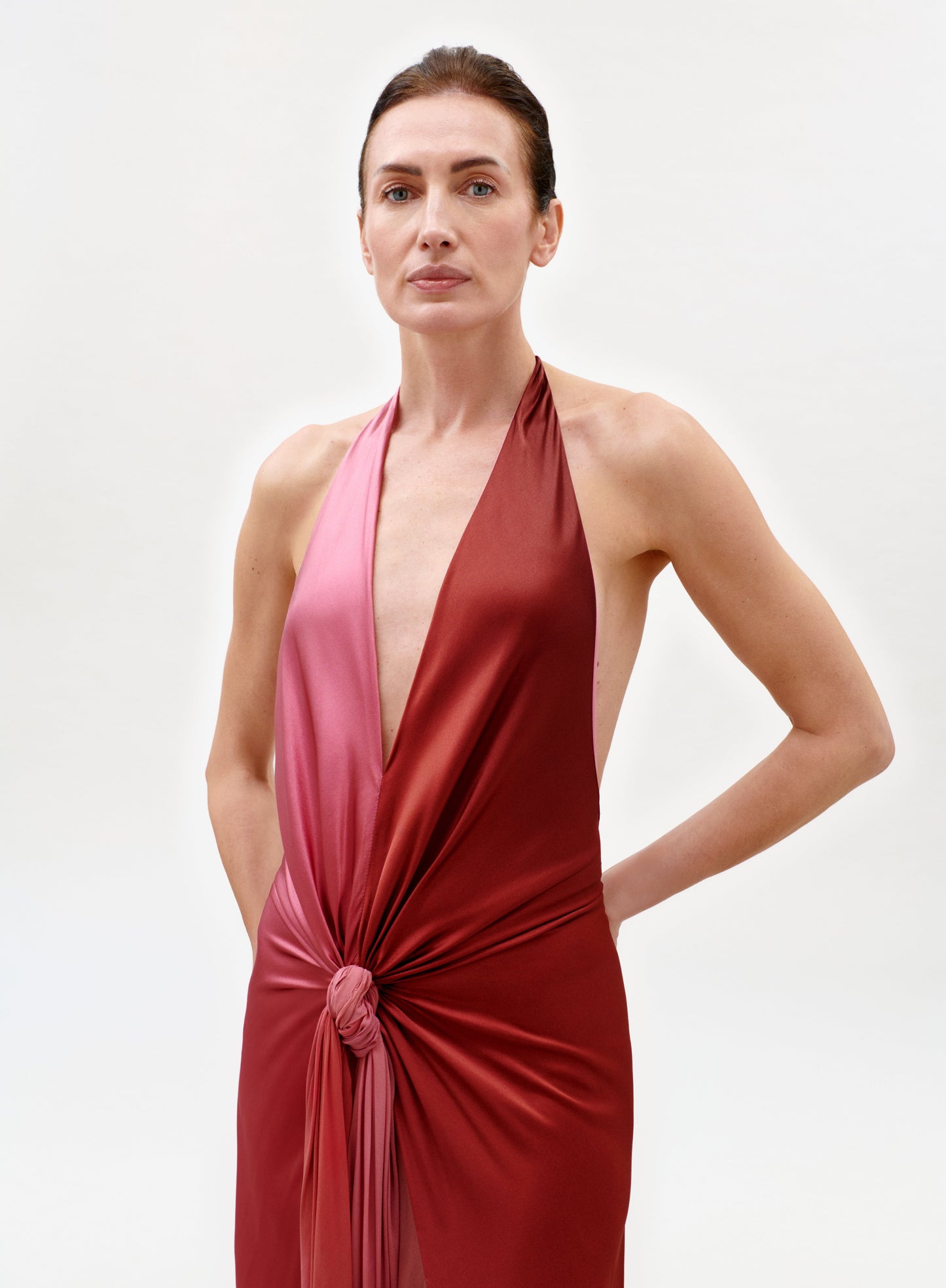 A woman with brown hair pulled back wears the Corsario Dress, a deep red and pink halter style with a front knot, standing with one hand in her pocket against a plain white background.