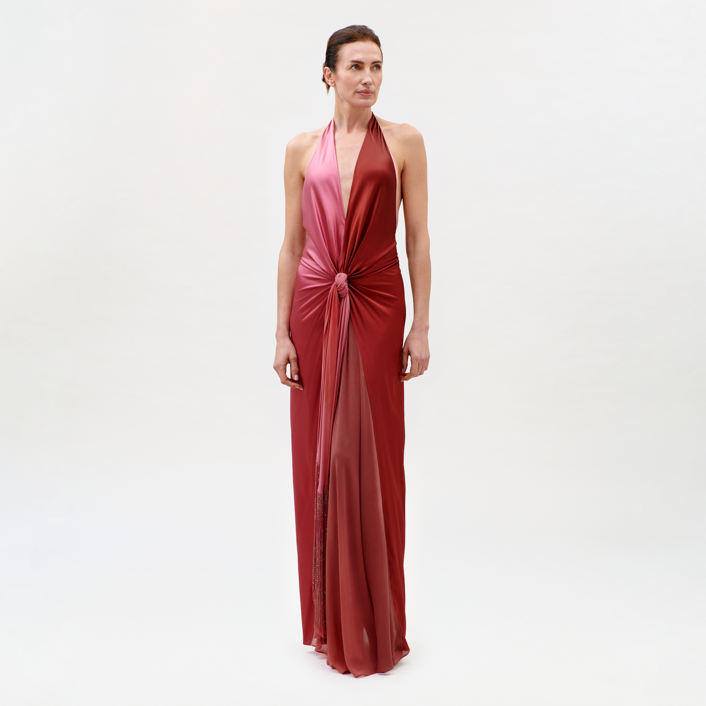 A woman stands against a light background, wearing the Corsario Dress—a sleeveless, deep rose halter gown with a knotted waist and long, flowing skirt. She gazes slightly to the side with a neutral expression.