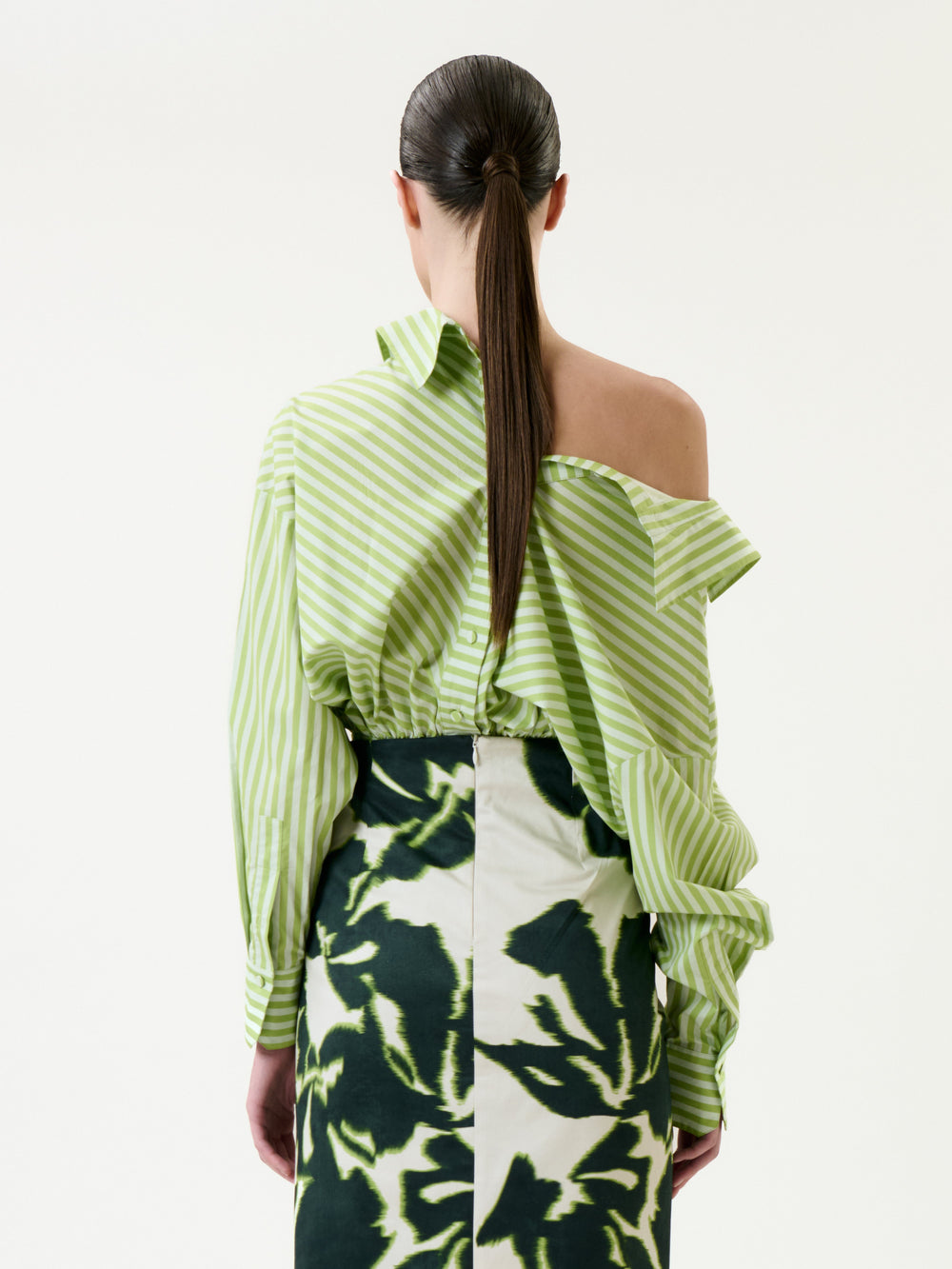 A person with long dark hair in a ponytail is shown from the back against a plain background, wearing the Skye Blouse—a light green striped shirt with an asymmetric neckline—paired with a green abstract patterned skirt.