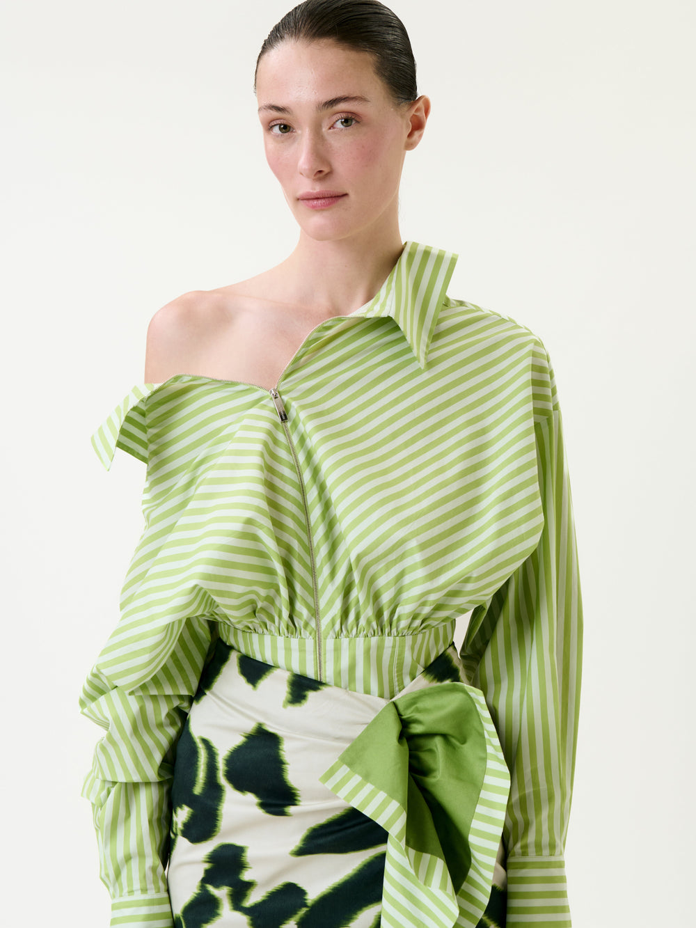 A person wearing the Skye Blouse—a light green and white striped off-shoulder top—paired with a skirt featuring a green abstract pattern and a large bow, standing against a plain background.