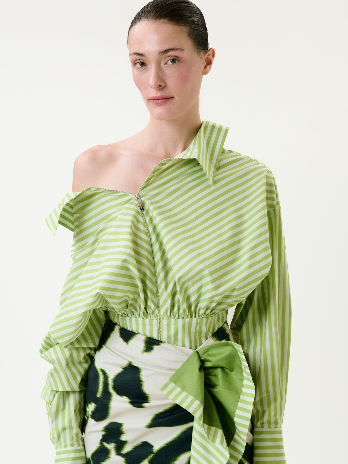 A person wearing the Skye Blouse—a light green and white striped off-shoulder top—paired with a skirt featuring a green abstract pattern and a large bow, standing against a plain background.