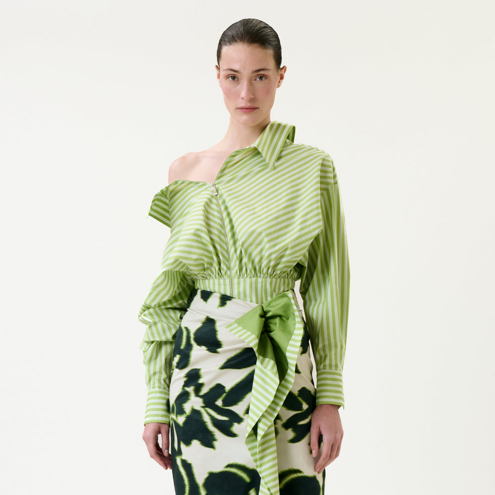 A woman wears the Skye Blouse, featuring green-and-white pinstripes, an asymmetrical cut, and ruffle details, paired with a floral-patterned skirt with a tied sash, standing against a plain white background.