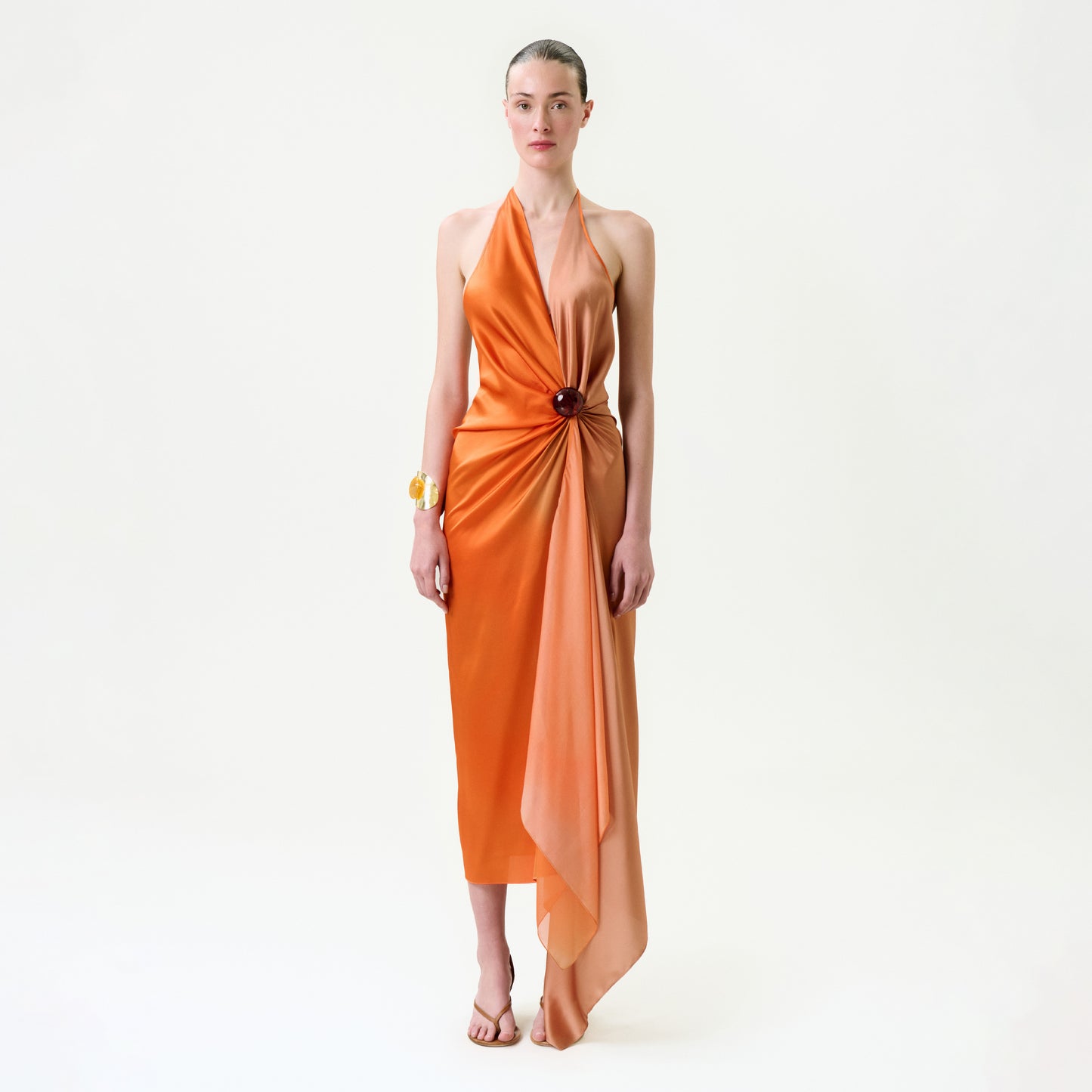 A woman poses against a white background wearing the Syndney Dress, an orange silk maxi with an asymmetrical hem and gathered waist draping, made from luxurious Italian fabric, styled with gold jewelry and nude sandals.