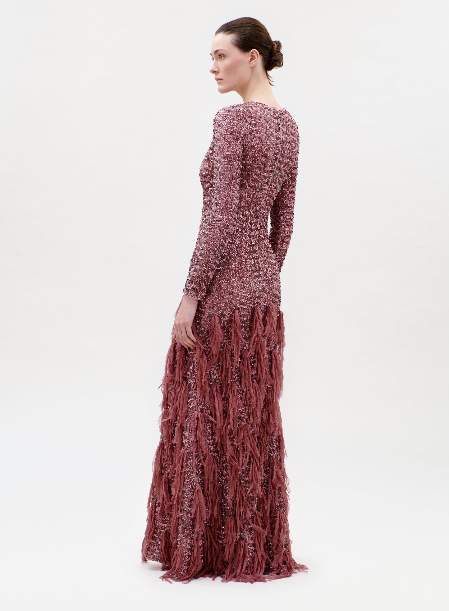 A woman stands in profile wearing the Bilal Dress, a long-sleeved, floor-length textured gown in dusty rose with fringe detailing from the waist down. The background is plain and light-colored.