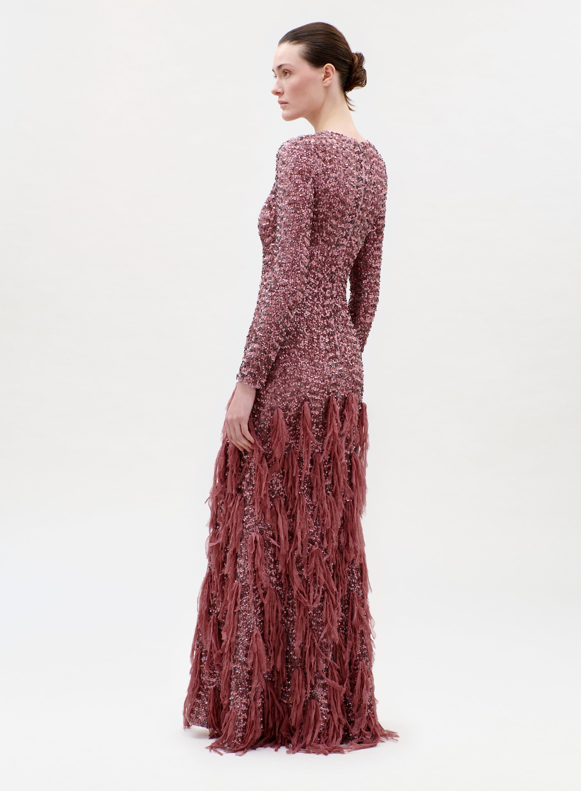 A woman stands in profile wearing the Bilal Dress, a long-sleeved, floor-length textured gown in dusty rose with fringe detailing from the waist down. The background is plain and light-colored.