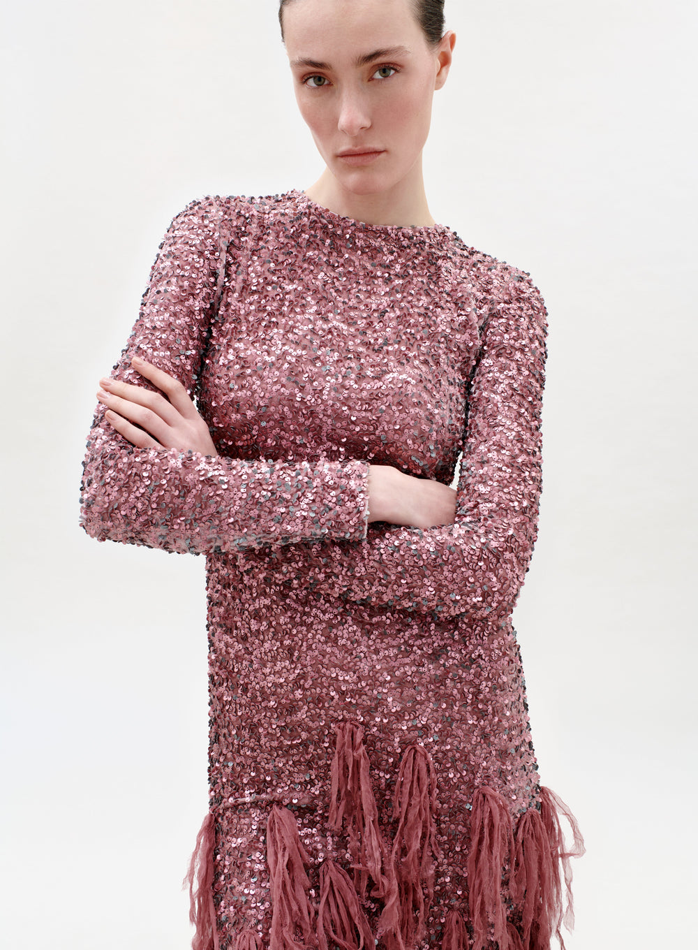A person with light skin and brown hair wears the Bilal Dress—a long-sleeved pink sequin dress with fringe details—standing arms crossed and neutral-faced against a plain white background.