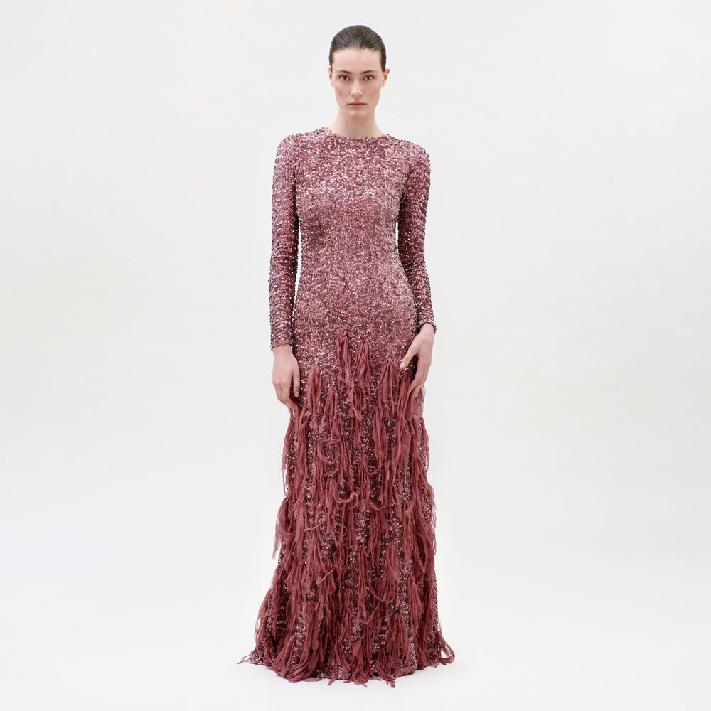 A woman faces the camera against a plain white background, wearing the Bilal Dress—a long-sleeved, floor-length pink evening gown featuring textured skirt detailing and a fitted bodice.