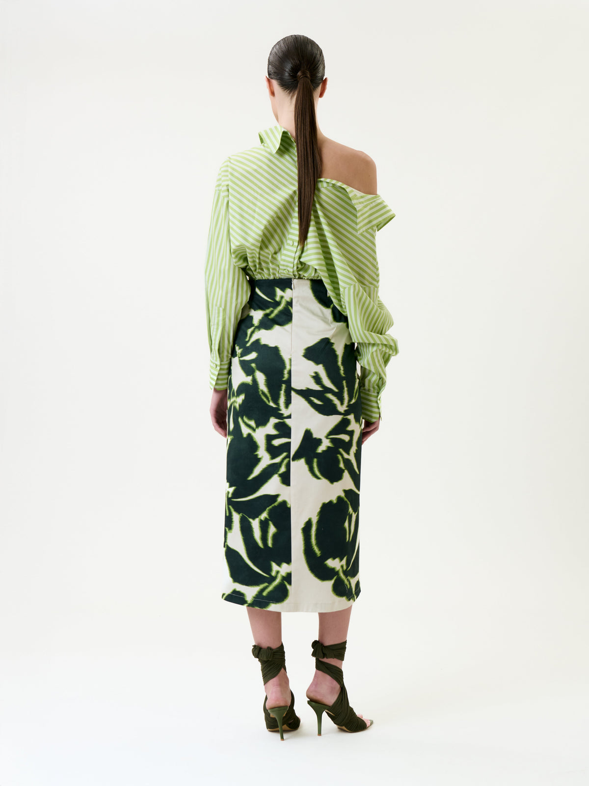 A woman faces away, wearing a green striped off-shoulder top paired with the Luana Skirt—a high-waisted piece featuring bold green and white abstract floral prints. Her hair is tied back, and she completes the look with dark green heeled sandals with ankle ties.