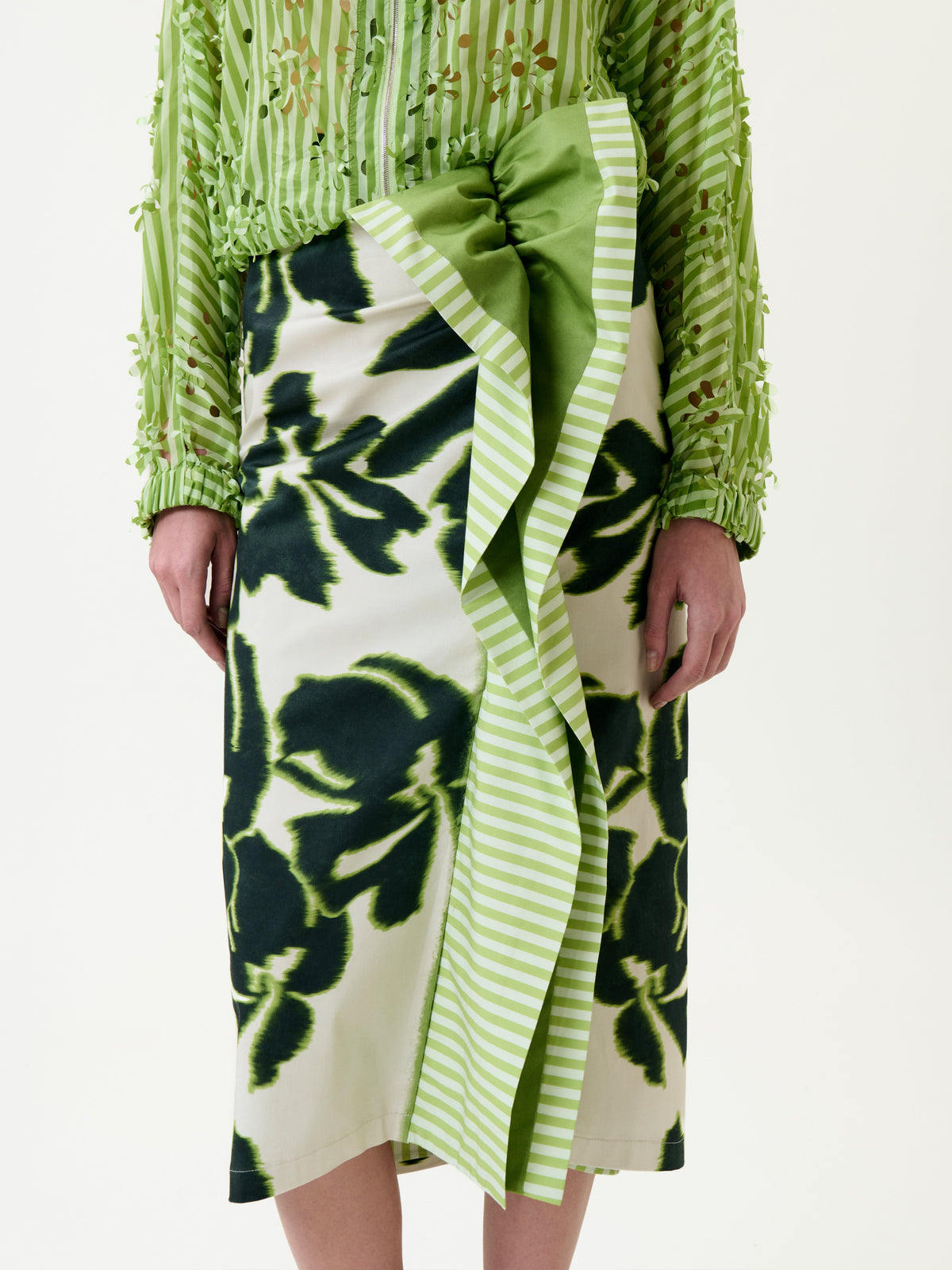 A person wears a green and white striped blouse with textured details and the Luana Skirt—a floral midi skirt featuring a bold green pattern and a large green-striped bow on the front.