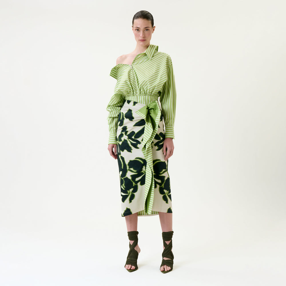 A woman stands against a white background wearing an asymmetrical green striped top, the Luana Skirt—a green and black floral wrap skirt—and olive green strappy heels.