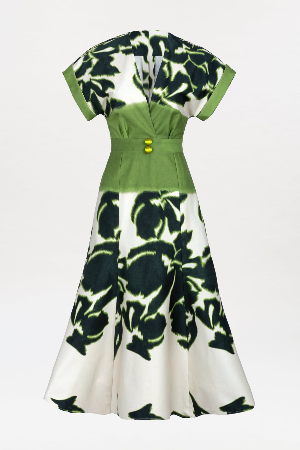 The Maida Dress is a knee-length design with short sleeves, a V-neckline, and a fitted waist. It features a green bodice adorned with yellow buttons and a white skirt decorated with bold, abstract black and green patterns.