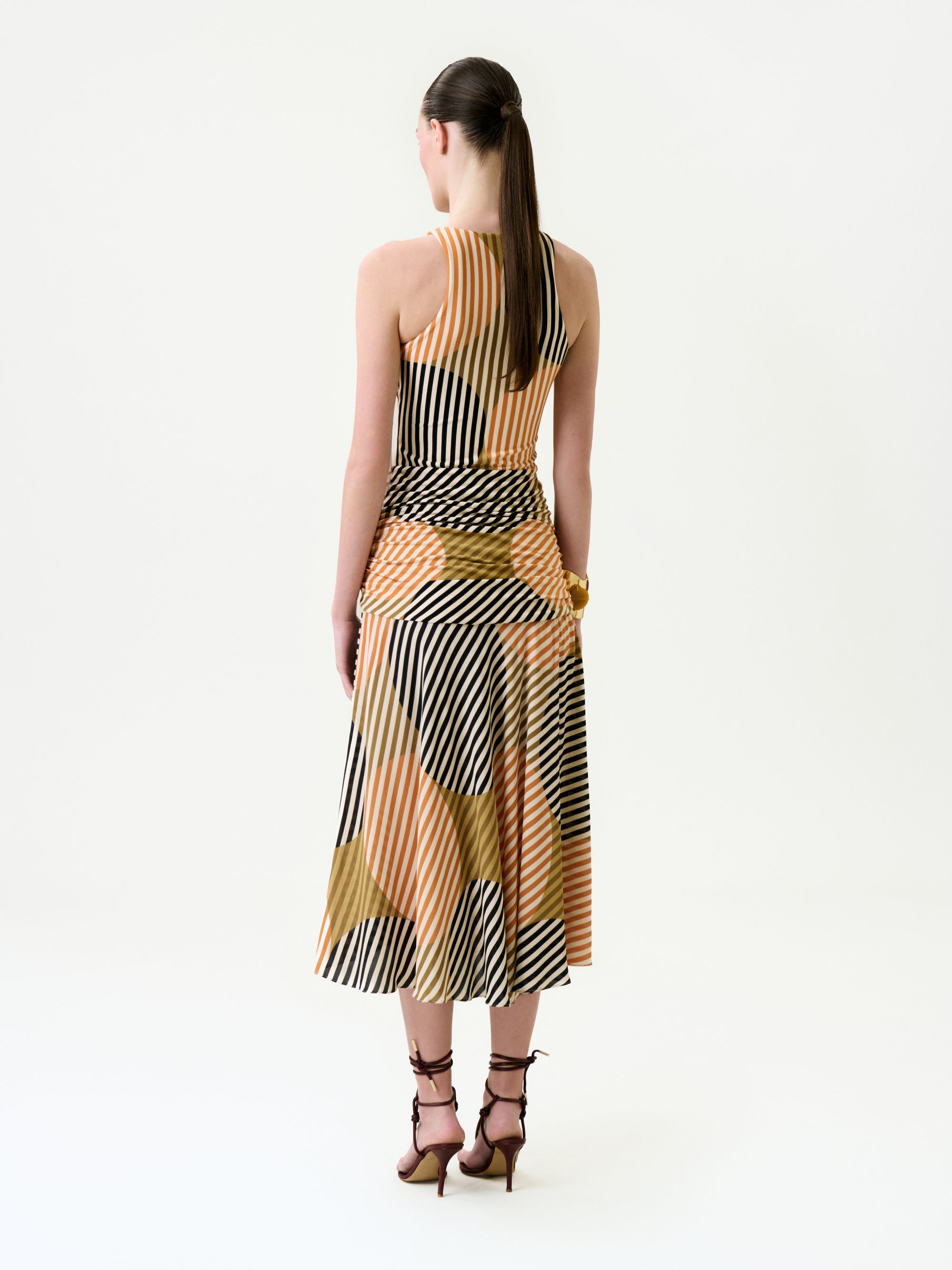 A woman with a long ponytail is standing, facing away, wearing the Malvina Dress—a sleeveless, calf-length midi crafted from luxurious Italian fabric with black, white, and tan geometric stripes—and brown strappy heels, all set against a plain white background.