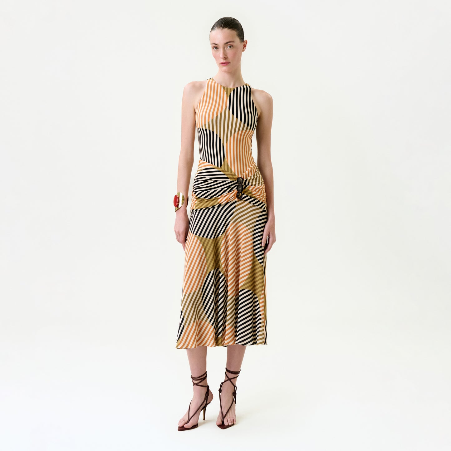 A woman poses against a plain white background wearing the Malvina Dress, a sleeveless, multicolored striped midi dress with a side knot crafted from luxurious Italian fabric, styled with strappy heels and a red cuff bracelet.
