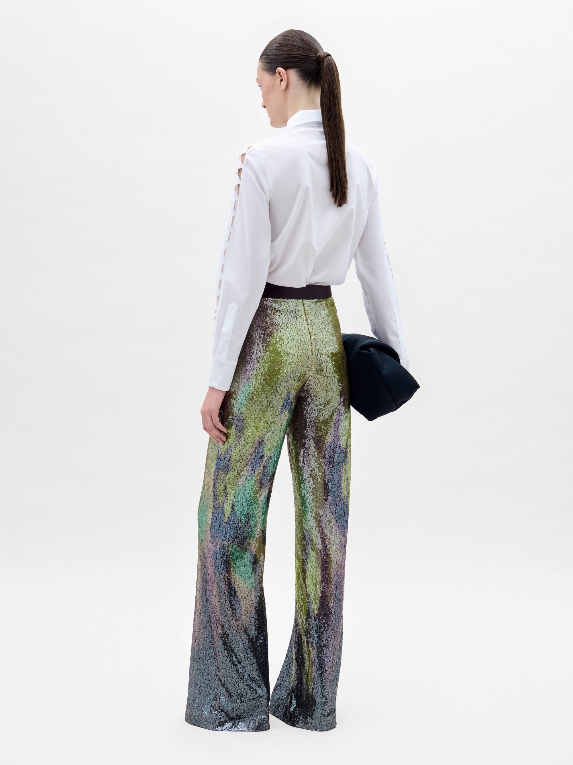 A person stands against a plain background wearing a white button-up shirt and the high-waisted, multicolored sequin Dalal Pant, holding a black clutch in their left hand.