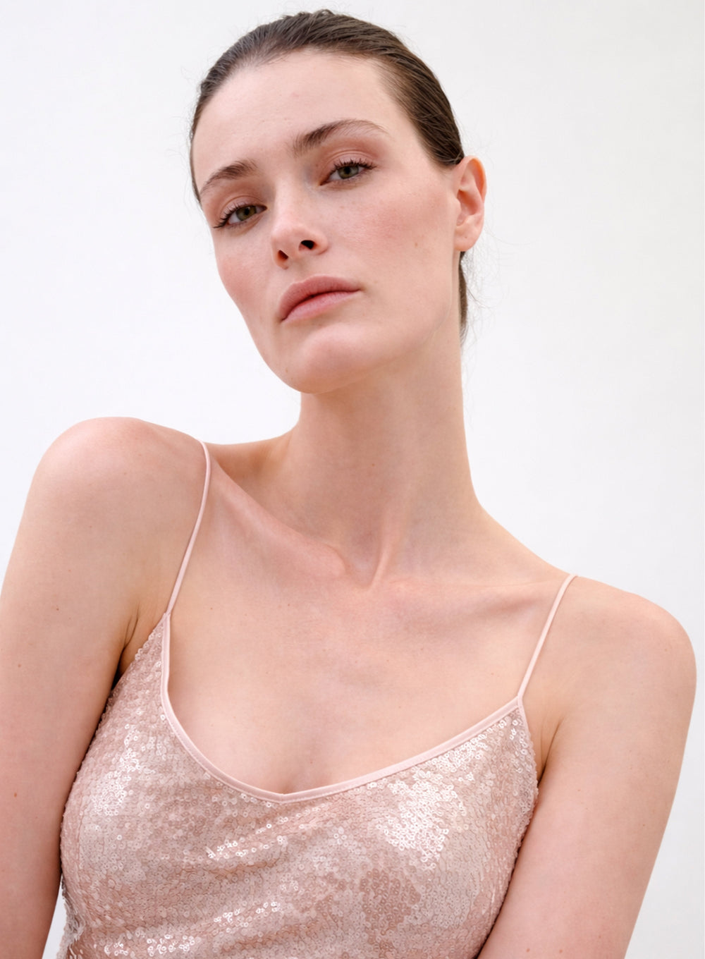 A woman with fair skin and brown hair looks at the camera with a neutral expression, wearing the Ibona Dress—a light pink, sequin-covered spaghetti strap top—against a plain white background.