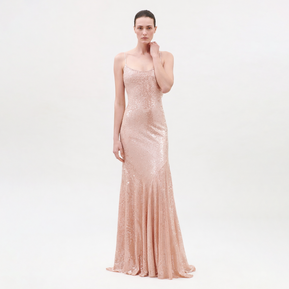 A woman with a neutral expression and pulled-back hair stands against a plain background wearing the Ibona Dress, a floor-length blush pink sequin gown with thin spaghetti straps and a form-fitting silhouette.