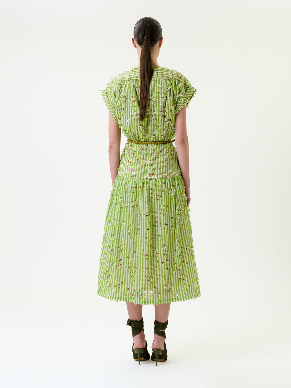A woman stands facing away, wearing the Ledicia Dress—a light green, textured, short-sleeved dress with a full skirt—paired with brown high-heeled sandals. Her straight brown hair is tied back in a low ponytail. The background is plain white.