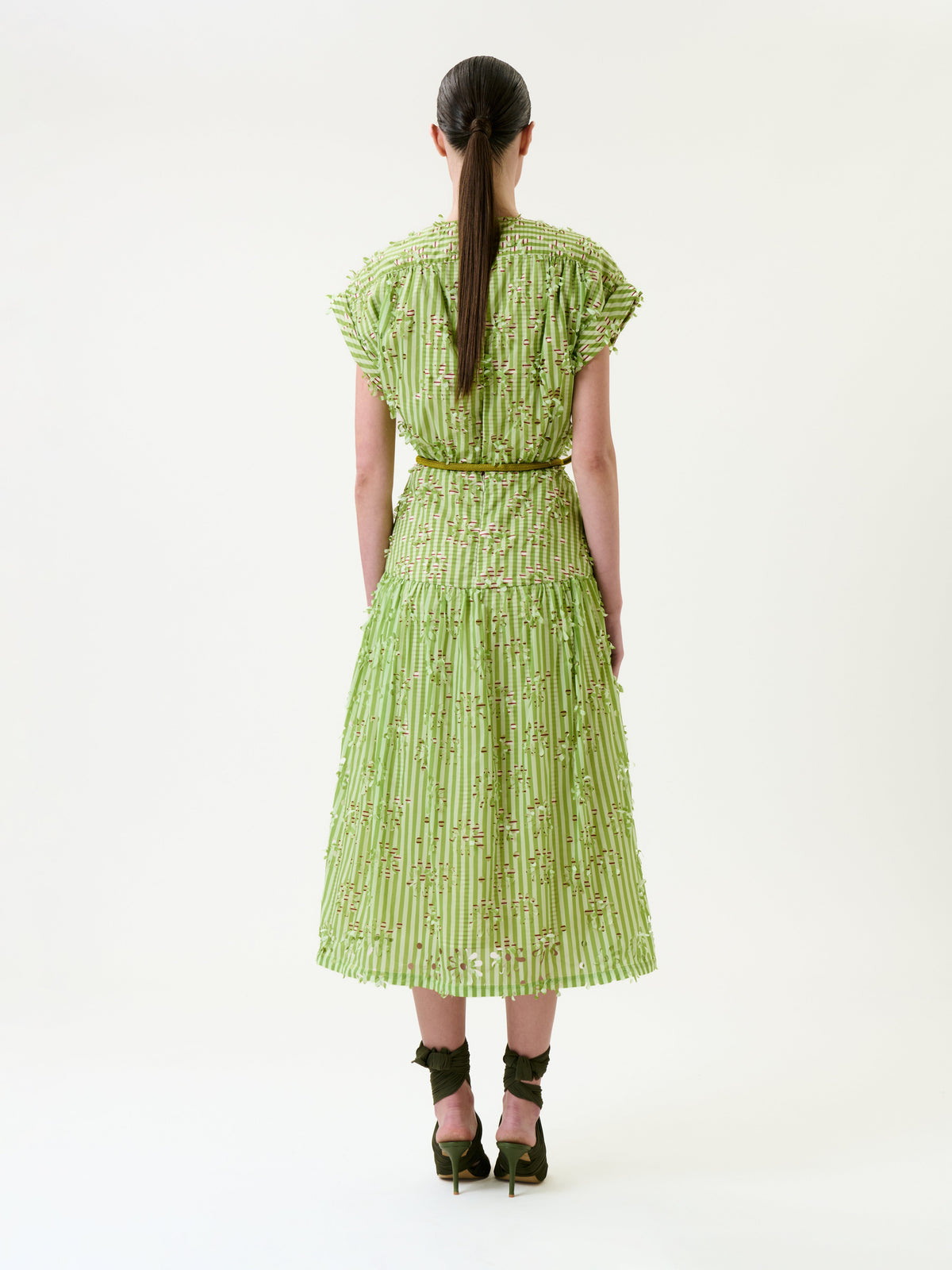 A woman stands facing away, wearing the Ledicia Dress—a light green, textured, short-sleeved dress with a full skirt—paired with brown high-heeled sandals. Her straight brown hair is tied back in a low ponytail. The background is plain white.