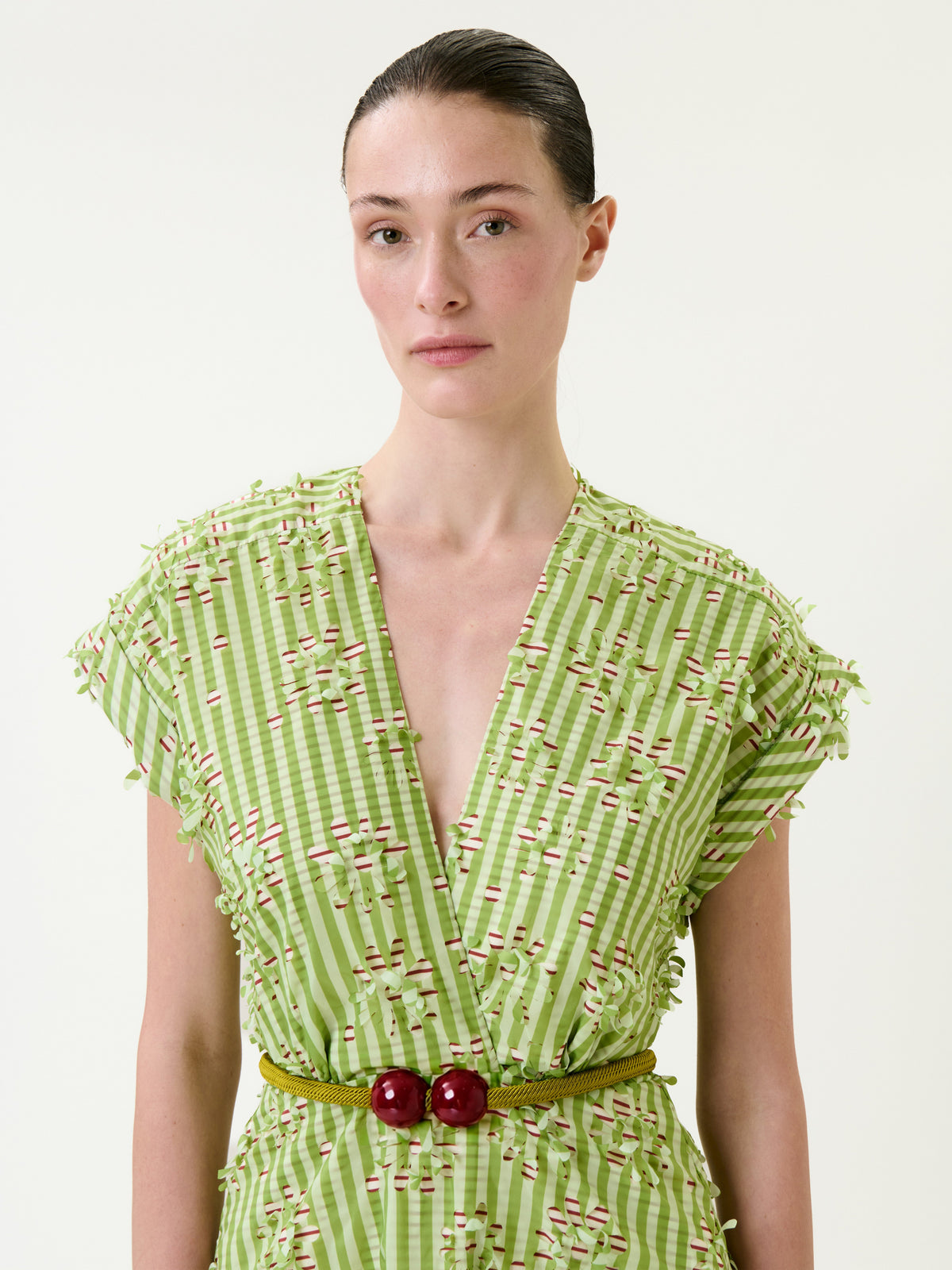 A woman with dark hair in a low bun wears the Ledicia Dress—a green checkered V-neck style with textured details and a braided belt adorned with two large red beads—standing against a plain light background.