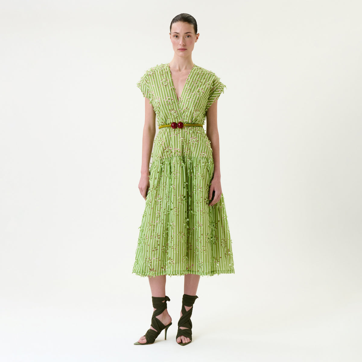A woman stands against a plain background, wearing the Ledicia Dress—a light green, pleated midi dress with a deep V-neck—paired with a red belt and dark green strappy heels. Her hair is pulled back.