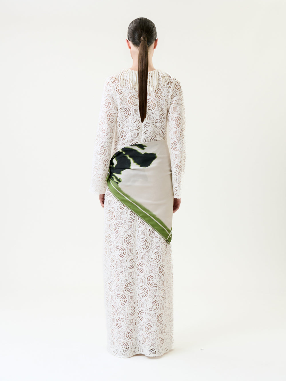 A person with long brown hair in a low ponytail wears the Vilma Dress, a floor-length white lace gown featuring a green and black patterned satin panel on the left hip, standing against a plain white background.