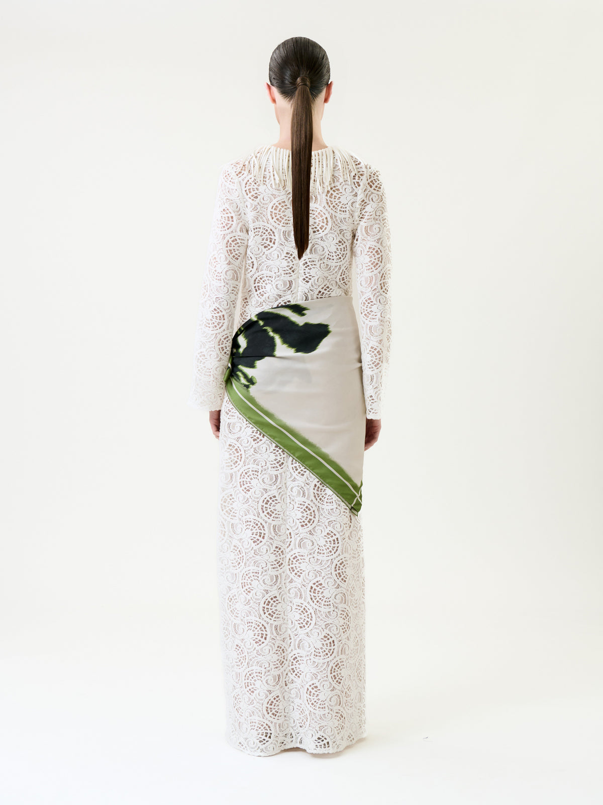 A person with long brown hair in a low ponytail wears the Vilma Dress, a floor-length white lace gown featuring a green and black patterned satin panel on the left hip, standing against a plain white background.