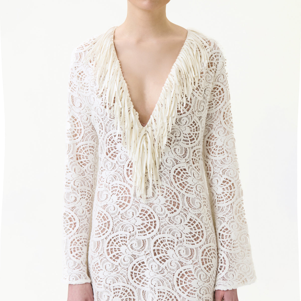 Wearing the Vilma Dress, a person showcases a white, long-sleeved lace design featuring a deep V-neckline adorned with long, cream-colored fringe detailing.