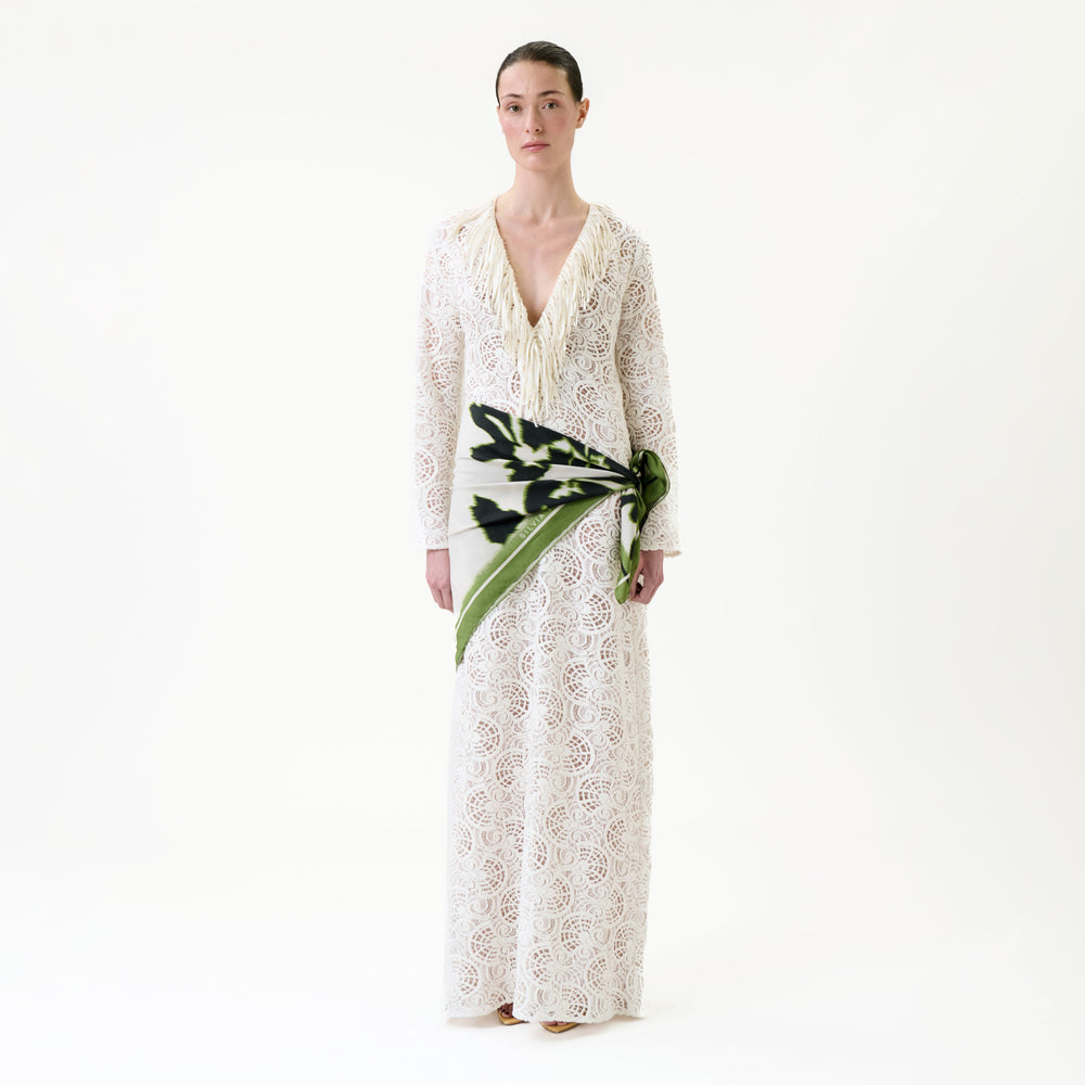 A woman stands against a white background, wearing the Vilma Dress—a long, white lace dress with a deep neckline—accented by a green and black patterned scarf tied around her waist.