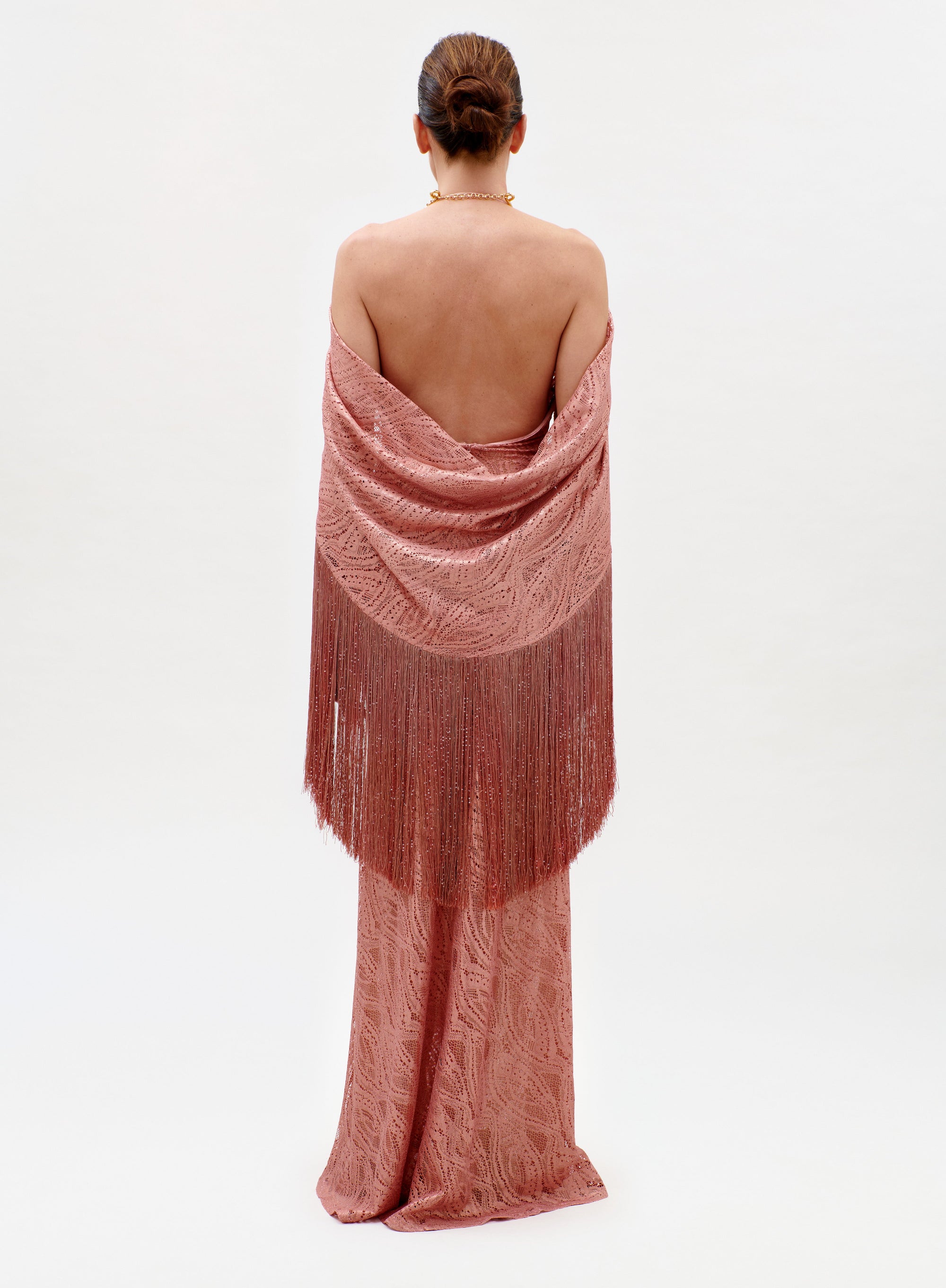 A person stands with their back to the camera, wearing the elegant, floor-length Habana Dress in textured pink, paired with a matching fringed shawl over bare shoulders against a plain white background.