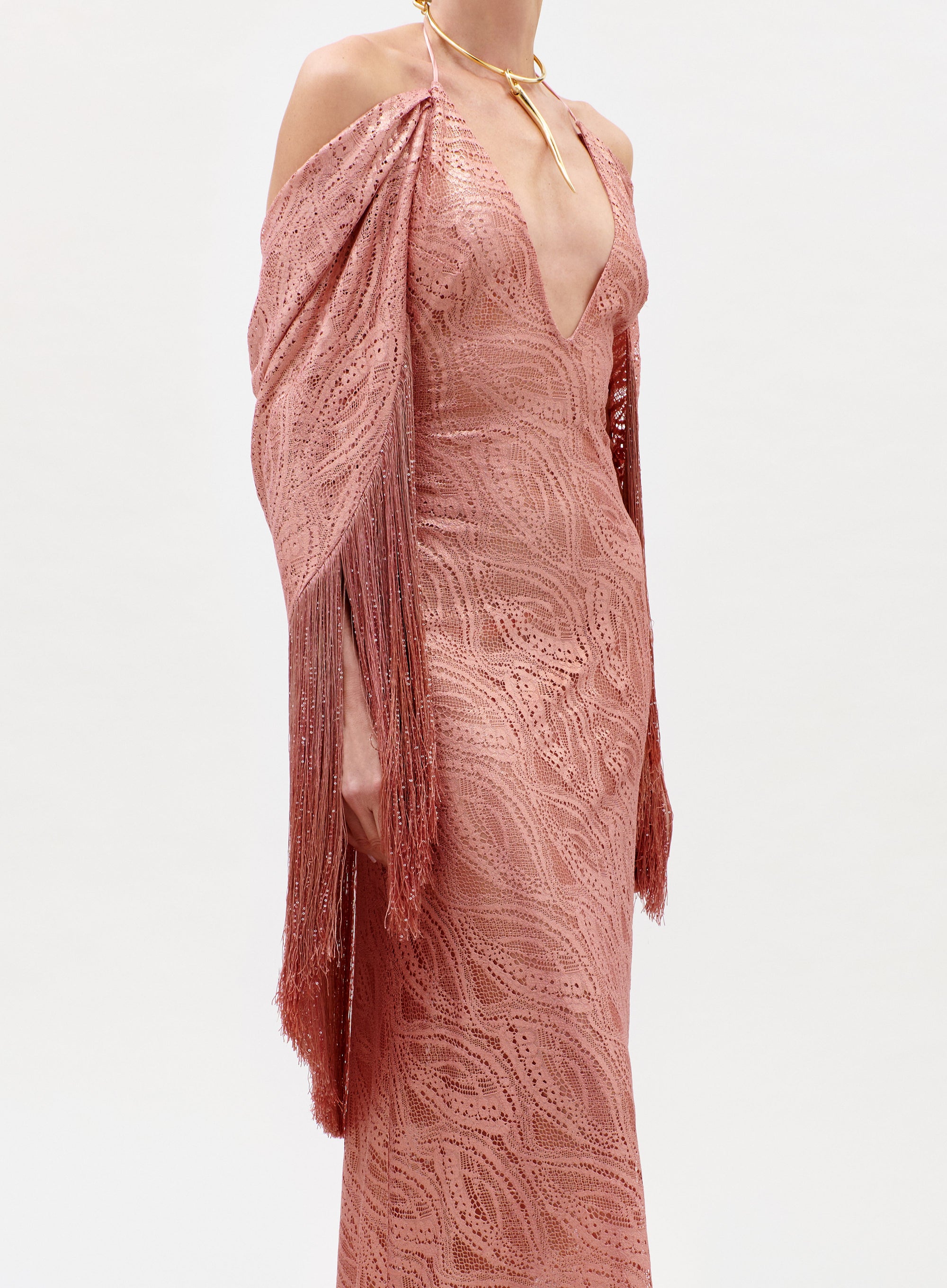A person wearing the Habana Dress—a pink lace gown with a deep V-neck, off-shoulder draped sleeves, fringed details—and a gold choker necklace, standing against a plain light background.