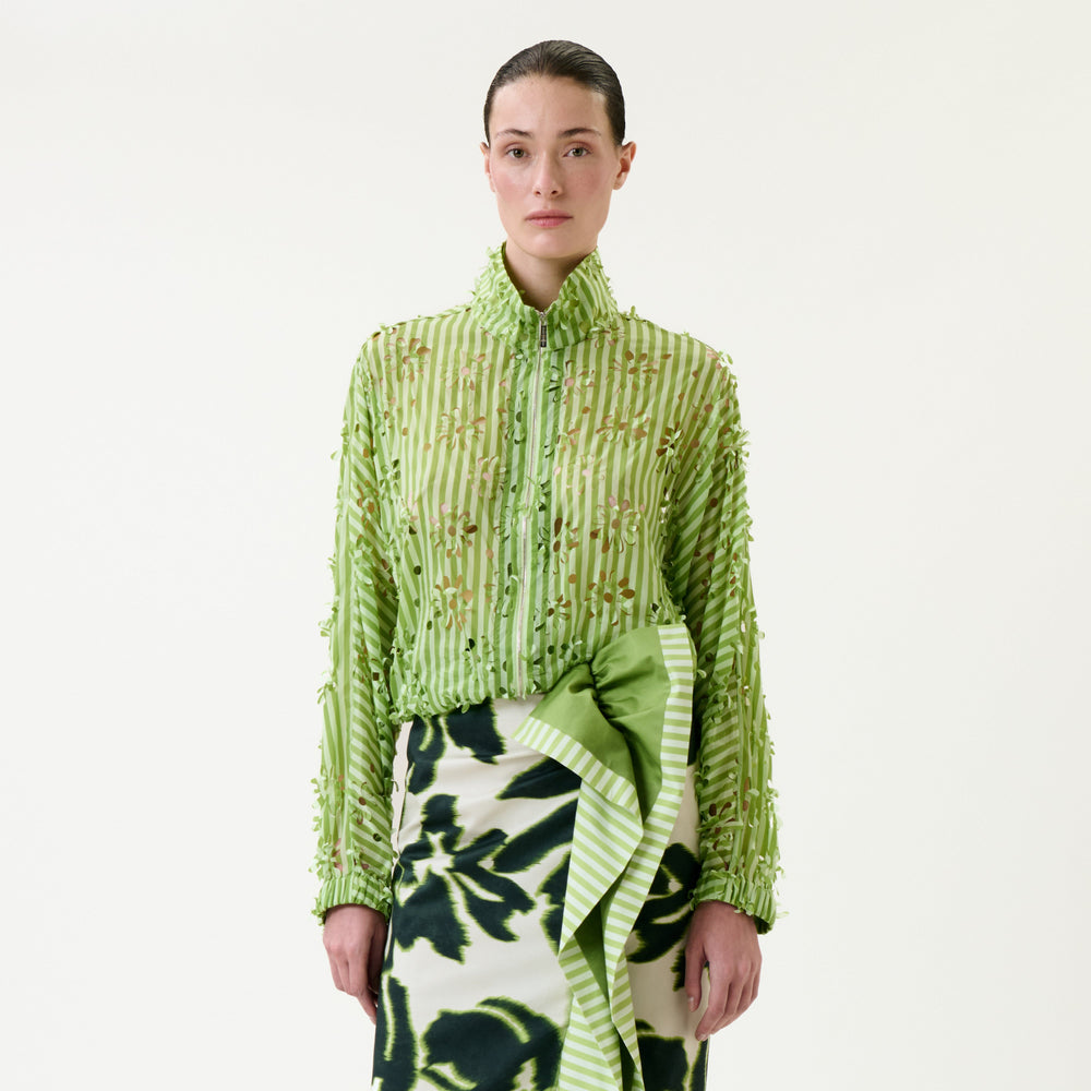 A woman stands facing forward, wearing the Randy Jacket—a green textured button-up shirt—paired with a white skirt featuring dark green floral patterns and a large green bow. The background is plain white.