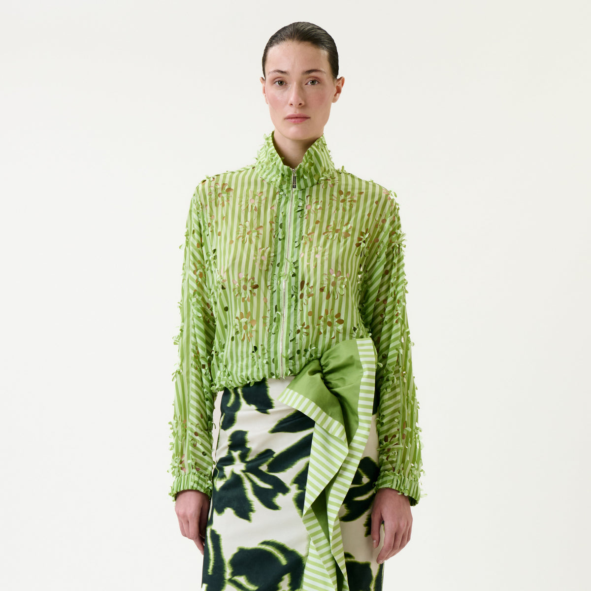 A woman stands facing forward, wearing the Randy Jacket—a green textured button-up shirt—paired with a white skirt featuring dark green floral patterns and a large green bow. The background is plain white.