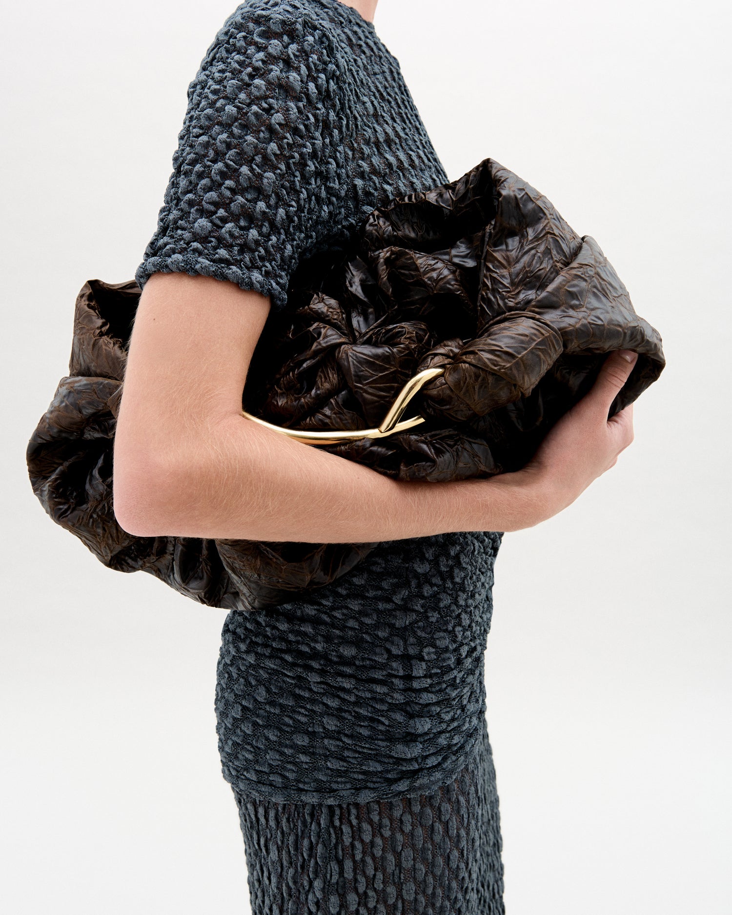 Person wearing a textured dark gray dress holding a brown leather clutch against a white background