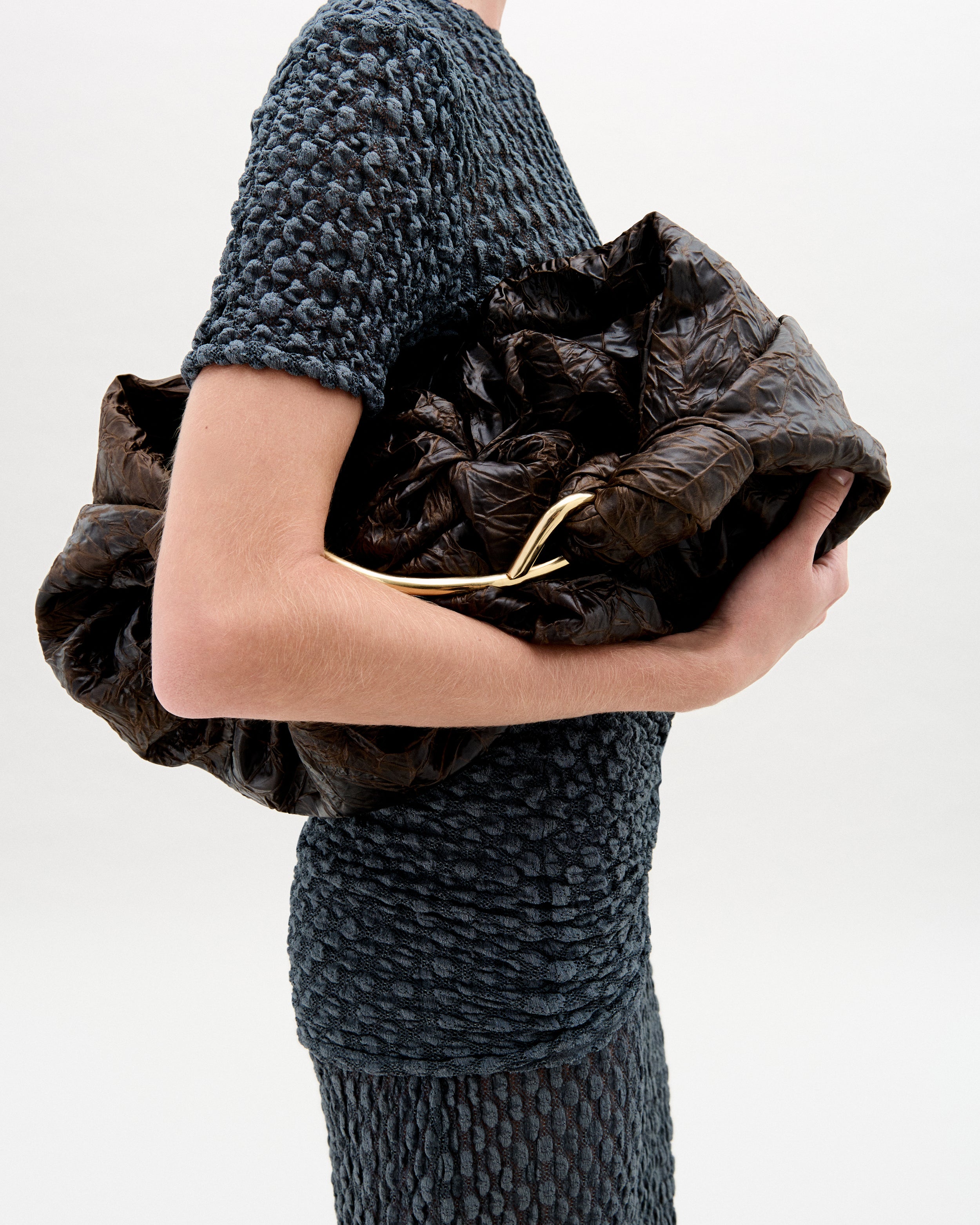 Person wearing a textured dark gray dress holding a brown leather clutch against a white background