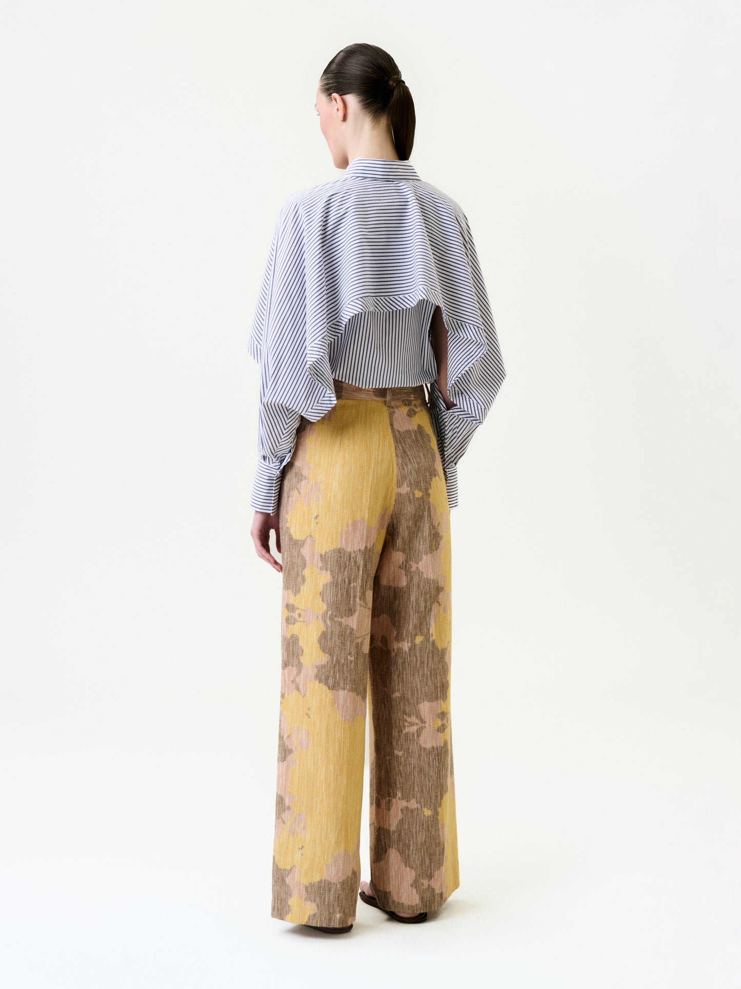A woman stands facing away, wearing wide-legged Emine Pants in a yellow and brown pattern with a relaxed Italian fabric fit, and a blue and white striped long-sleeve layered-back top, against a plain white background.