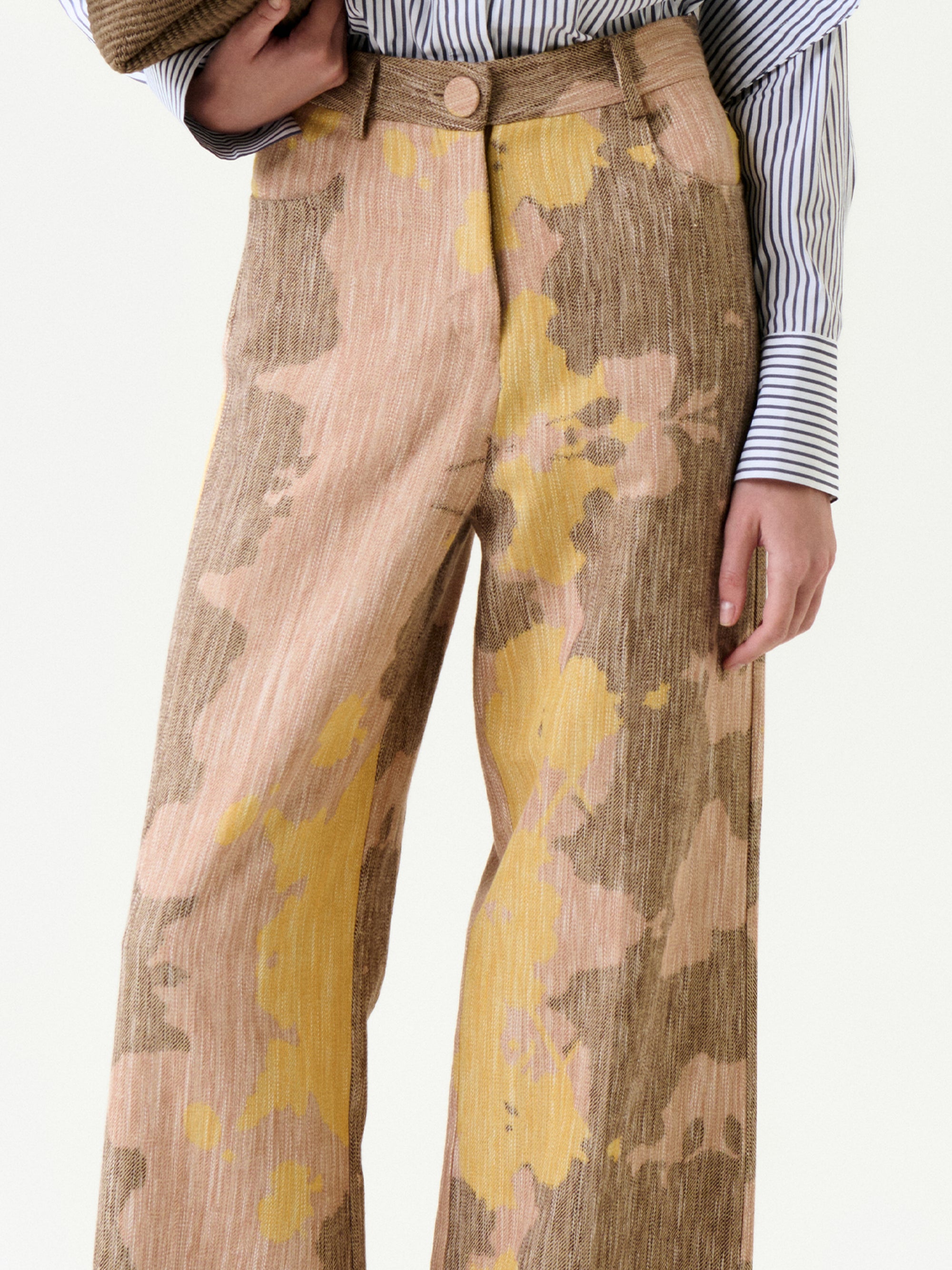 A person wears the Emine Pant, featuring a high-waisted, wide-leg relaxed fit with a yellow and brown abstract pattern, paired with a blue and white striped shirt. Only the torso and legs are visible.
