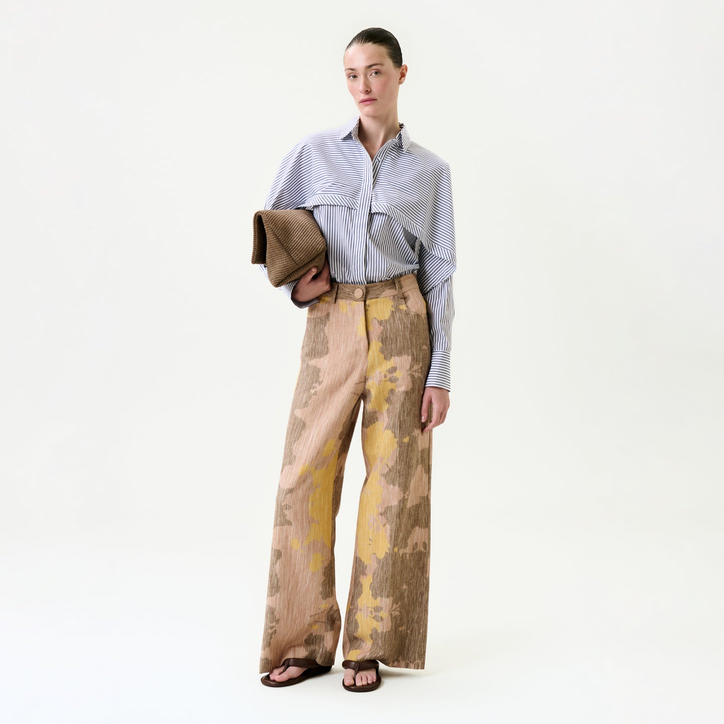 A woman stands against a plain background wearing a blue striped button-up shirt crafted from Italian fabric, the Emine Pant in beige and yellow, black sandals, and holding a brown textured clutch.