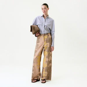 A woman stands against a plain background wearing a blue striped button-up shirt crafted from Italian fabric, the Emine Pant in beige and yellow, black sandals, and holding a brown textured clutch.