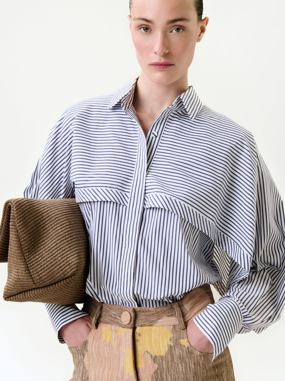 Wearing the Nira Shirt, a relaxed fit blue and white striped button-up, paired with brown pants featuring yellow accents, a person holds a textured brown clutch under one arm against a plain white background.