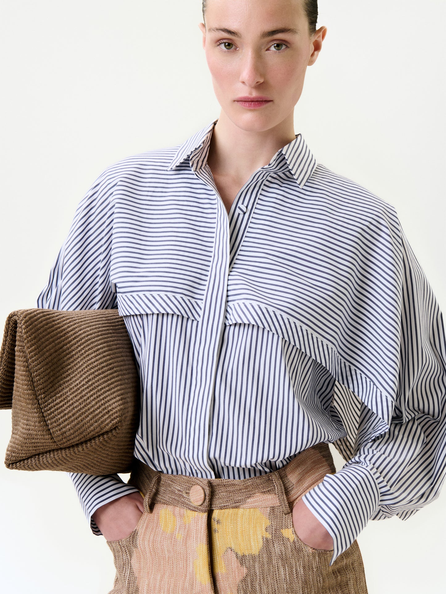 Wearing the Nira Shirt, a relaxed fit blue and white striped button-up, paired with brown pants featuring yellow accents, a person holds a textured brown clutch under one arm against a plain white background.