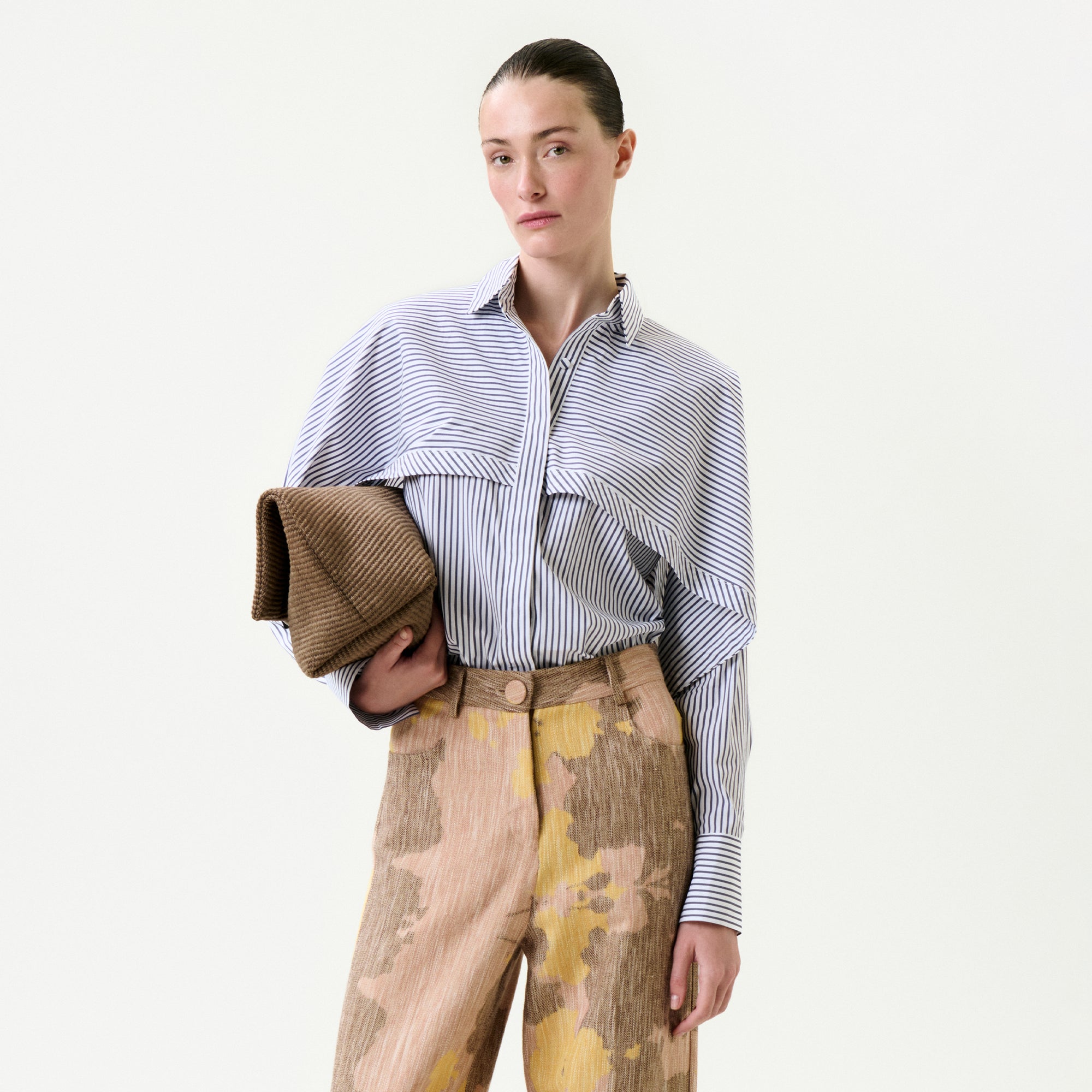 A person with slicked-back hair wears the relaxed fit Nira Shirt in blue and white stripes, paired with high-waisted beige and yellow patterned pants, holding a textured brown clutch against a plain light background.