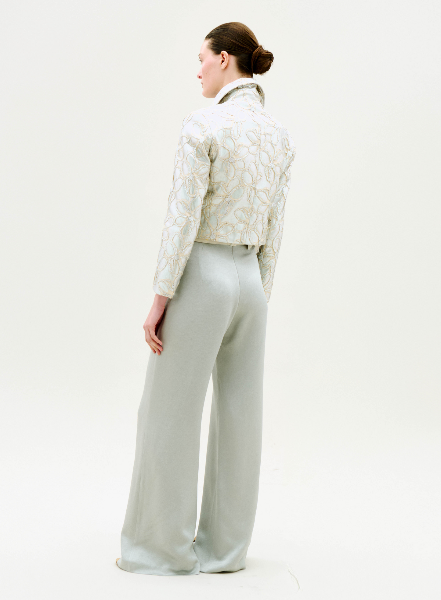 A woman stands facing away, wearing the Perla Jacket and high-waisted, wide-leg light gray pants. Her hair is in a neat bun against a plain white background.