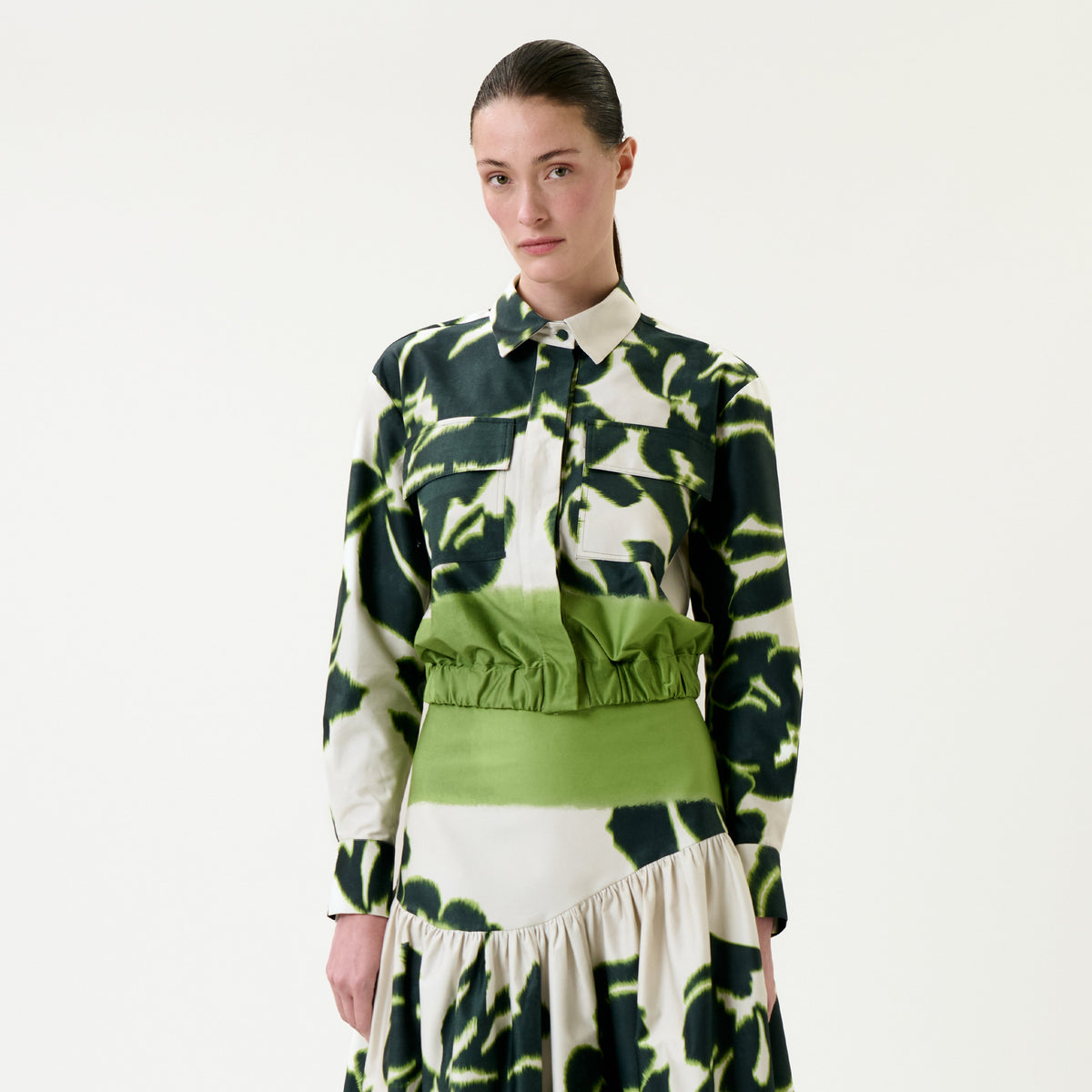 A woman stands against a plain white background, wearing the Soleil Jacket, which features a green and cream pattern with a high waist detail and long sleeves. She has straight brown hair pulled back and a neutral expression.