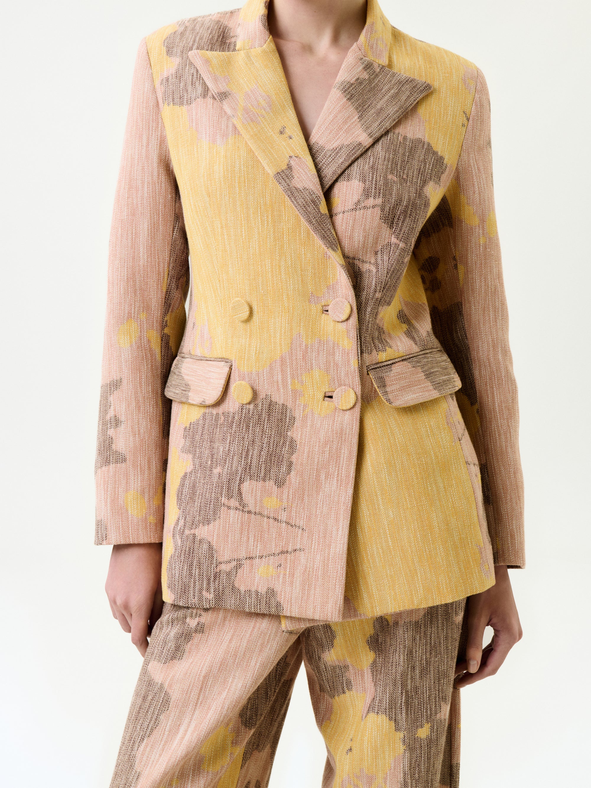 A person wearing the Frankie Jacket, a textured double-breasted suit in light pink, yellow, and brown abstract Italian fabric with flap pockets and wide lapels. The true-to-size jacket is paired with matching pants for a complete look.