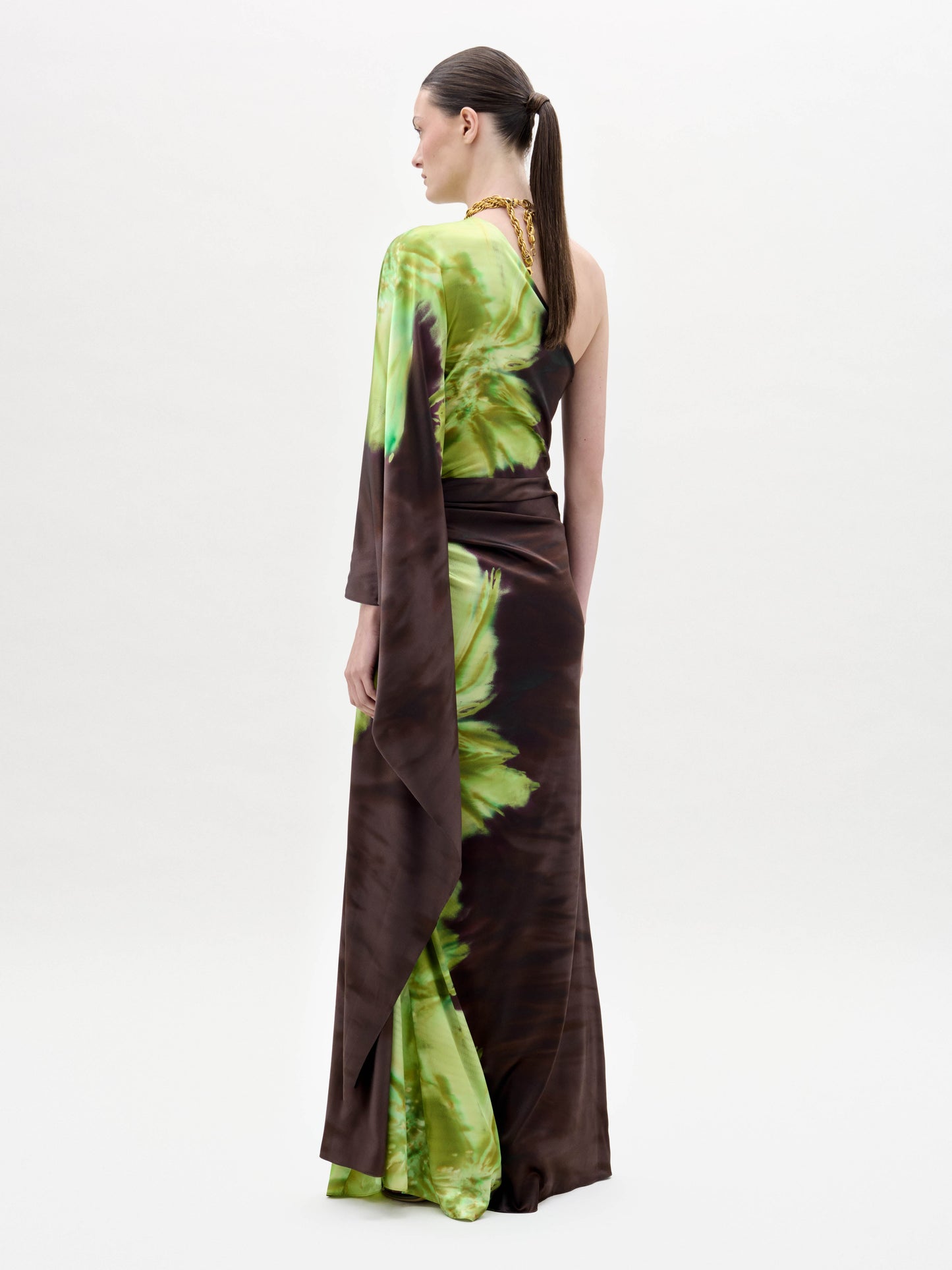 A person models the Afrodita Dress—a one-shoulder, floor-length gown with a green and brown tie-dye print—paired with gold necklaces, against a plain white background.