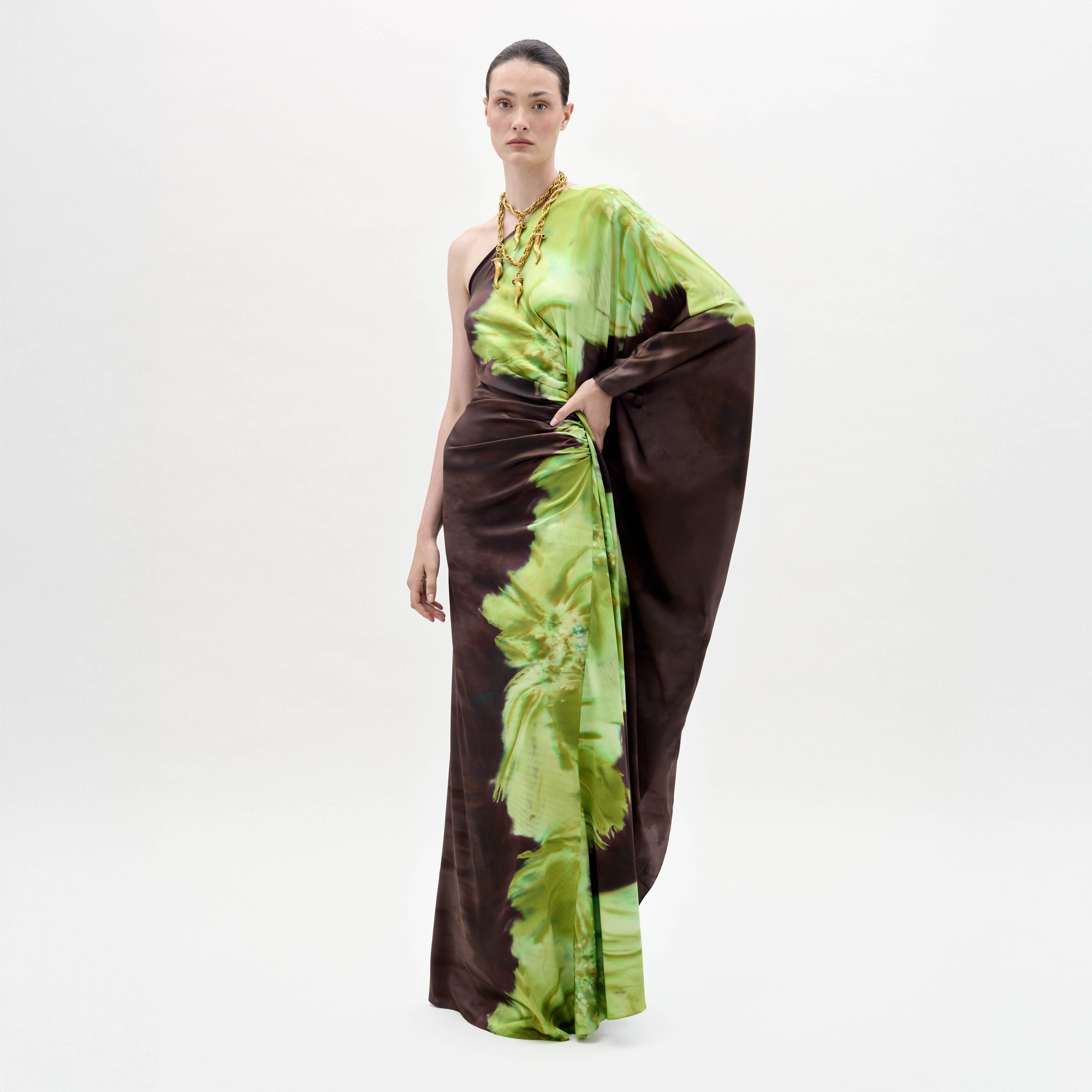 A person models the Afrodita Dress—a one-shoulder, floor-length gown with a green and brown tie-dye print—paired with gold necklaces, against a plain white background.