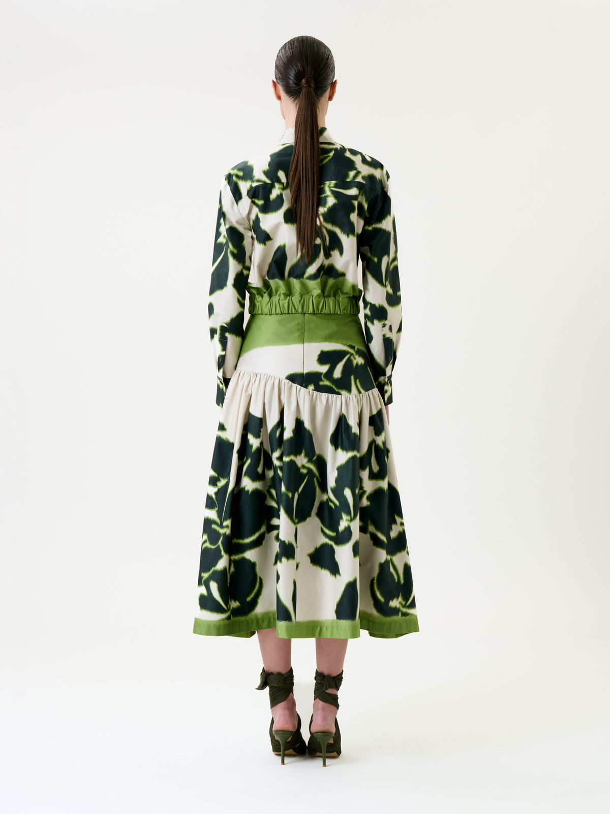 A woman stands facing away, wearing the Libby Skirt—a long-sleeve, green and white patterned skirt with a wide green waistband and hem—paired with green heels featuring ribbon ties. Her hair is tied back in a low ponytail.